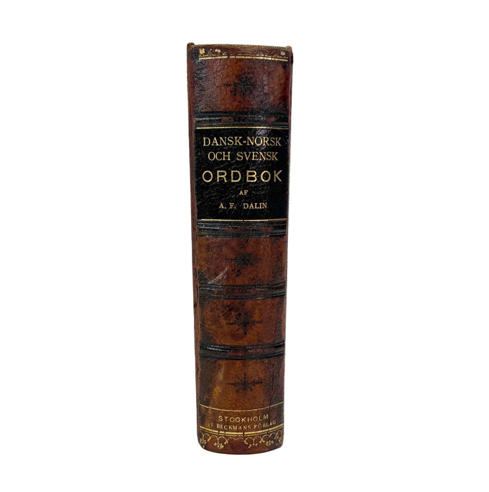 A vintage leather-bound dictionary standing upright on a white studio background. The book spine is dark brown with gold lettering reading 'DANSK-NORSK OCH SVENSK ORDBOK' and 'A.
