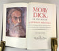 An open book showing the title page of 'Moby Dick: Or, the Whale' by Herman Melville, with a painted portrait of Moby Dick on the left page and red and black typography on an off-white page. The book is held open by two gold-toned bookends on a light gray surface.