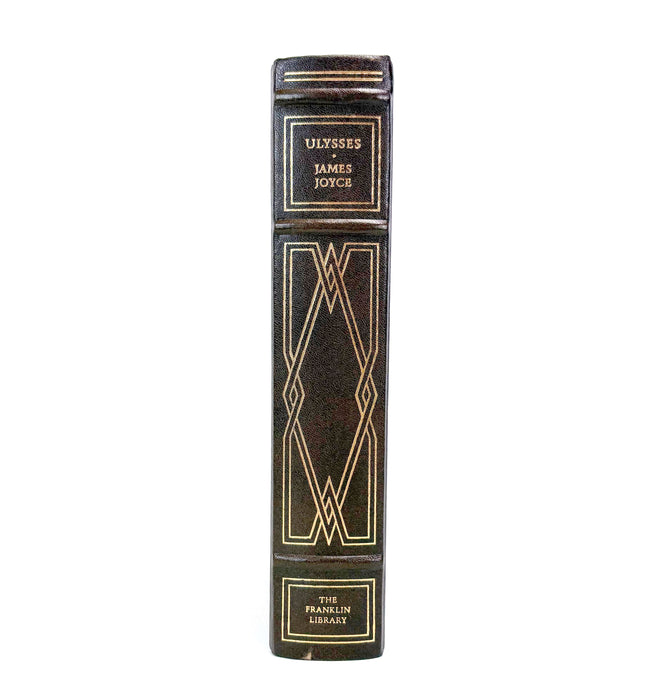 A close-up of a book spine for 'Ulysses' by James Joyce, featuring a dark brown or black leather-like cover with gold decorative geometric patterns and gold lettering, isolated on a white background.